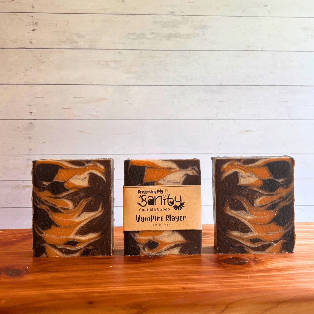 Three bars of Vampire Slayer goat milk soap displayed in a row on a cedar bath board with a white paneled background. Two bars are displayed to show the Taiwan swirl pattern of black, brown, white, and orange in this cozy fall soap. The third bar of soap has a label on it.