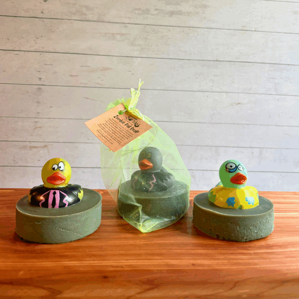 Three bars of Zombie Pals goat milk soap sitting on a cedar bath board. The soap bars are an olive green color and each bar of soap has a rubber zombie toy lightly embedded into the top. In the photo, one is pictured in a bag to show packaging, while the other two are unpackaged to show detail.