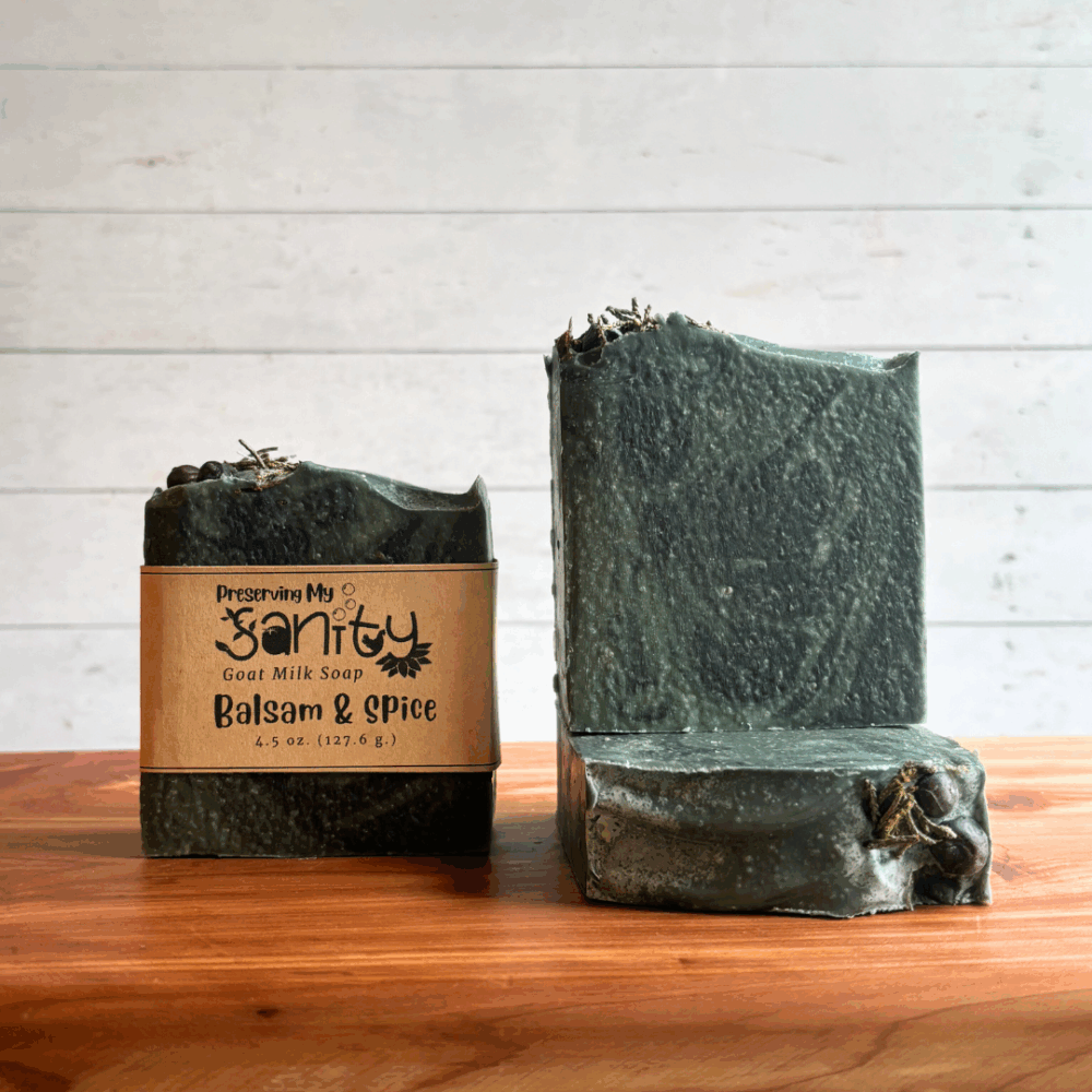 This Balsam & Spice goat milk soap is part of the holiday collection. It's dark green with black swirls and has spruce tips and juniper berries decorating the top. The photo shows three bars of soap of varying angles, one bar has a brand logo on it.