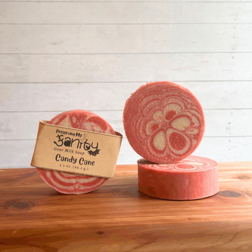 Three bars of round candy cane soap pictured on a cedar bath board with a white panel background. The bars of soap are red and white in a fun kaleidescope pattern. One soap bar features a label, the other two are bare.
