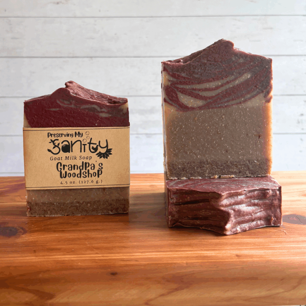 Three bars of Grandpa's Woodshop goat milk soap, pictured to show the brown textured bottom and swirled red and uncolored top. Picutred on a cedar bath board with a white paneled background.