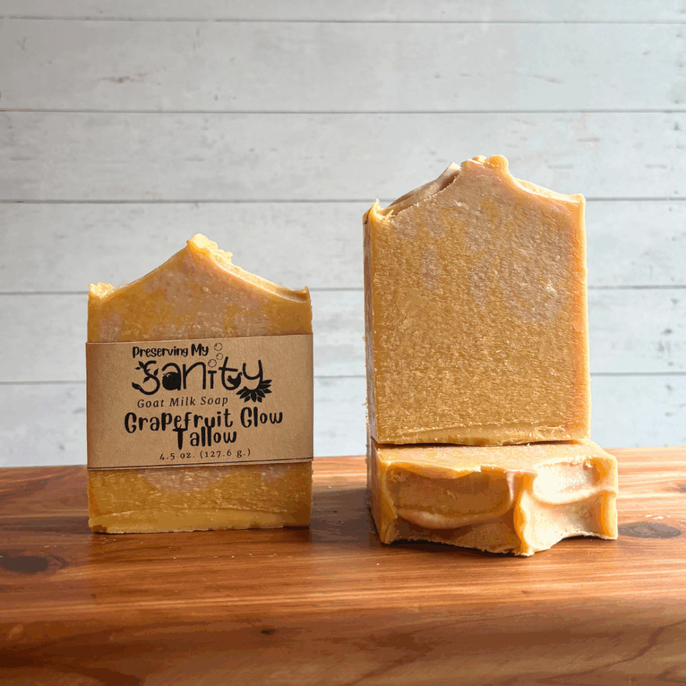 Three bars of Grapefruit Glow tallow soap pictured on a cedar bath board with white paneled background. Soap is made with goat milk sourced from local farmers, and the photo shows a few angles of the soap to display detail and label.