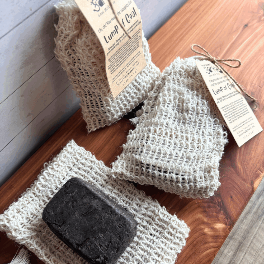 Alternate view of three lump of coal goat milk soap bars with reusable sisal soap saver bags with gift tags, one out of the bag to see soap bar detail.