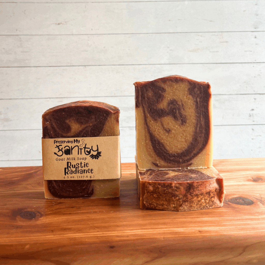 Three bars of Rustic Radiance goat milk soap, made with natural plant-based colorants and orange and patchouli essential oils. The soap is pictured to display different angles, and one bar has a label on it. Bars are sitting on a cedar bath board with a white paneled background.