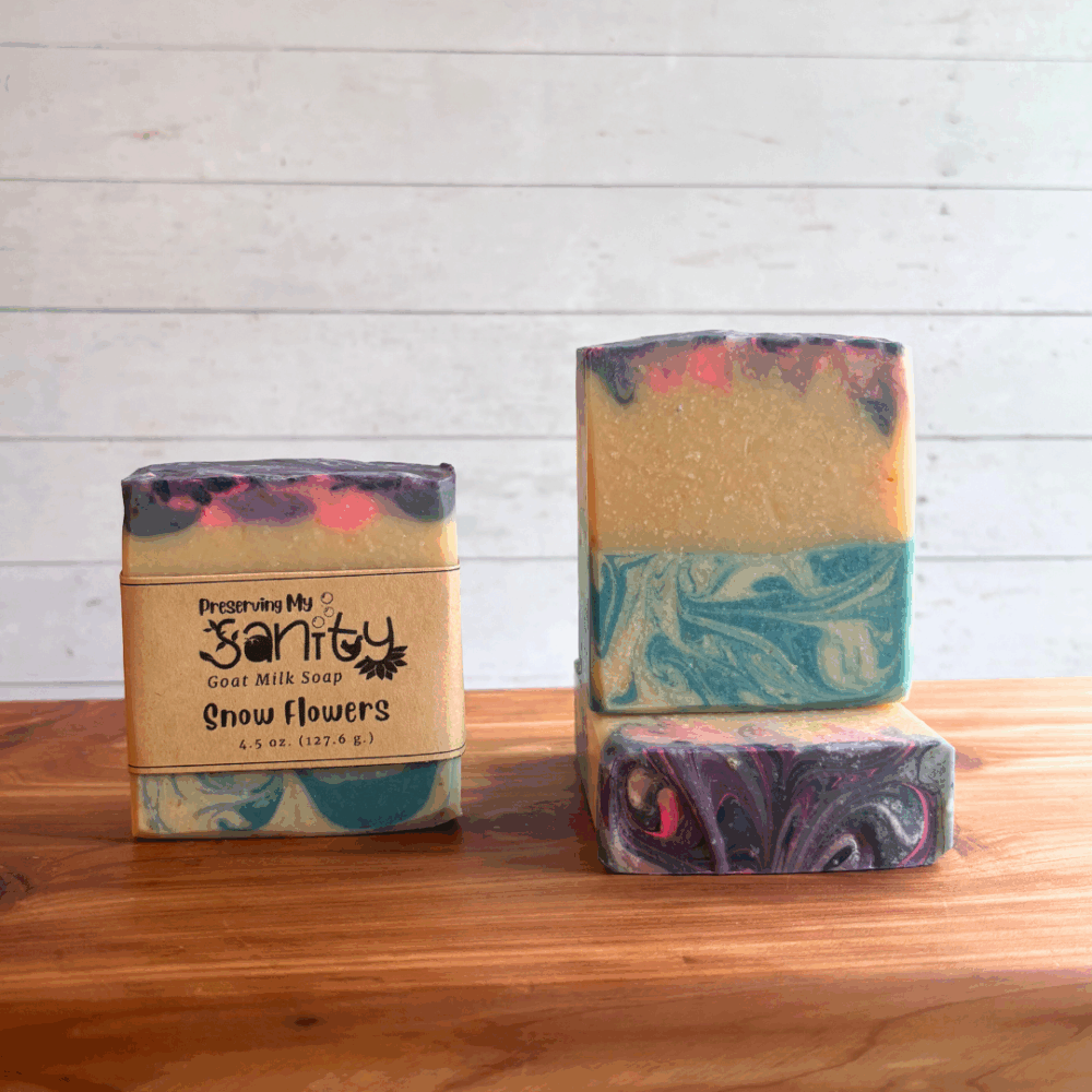 Three bars of Snow Flowers goat milk soap, pictured on a cedar bath board at varying angles to show different views of the soap. One bar has a label on it, the other two are bare. Bars are sitting on a cedar bath board with a white paneled background behind it.