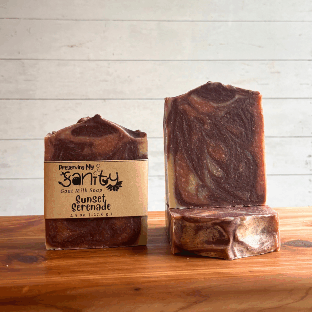 Three bars of Sunset Serenade goat milk soap situated on a cedar bath board with a white paneled background. Soap is made with an essential oil blend, natural plant-based colorants, and locally sourced goat milk.