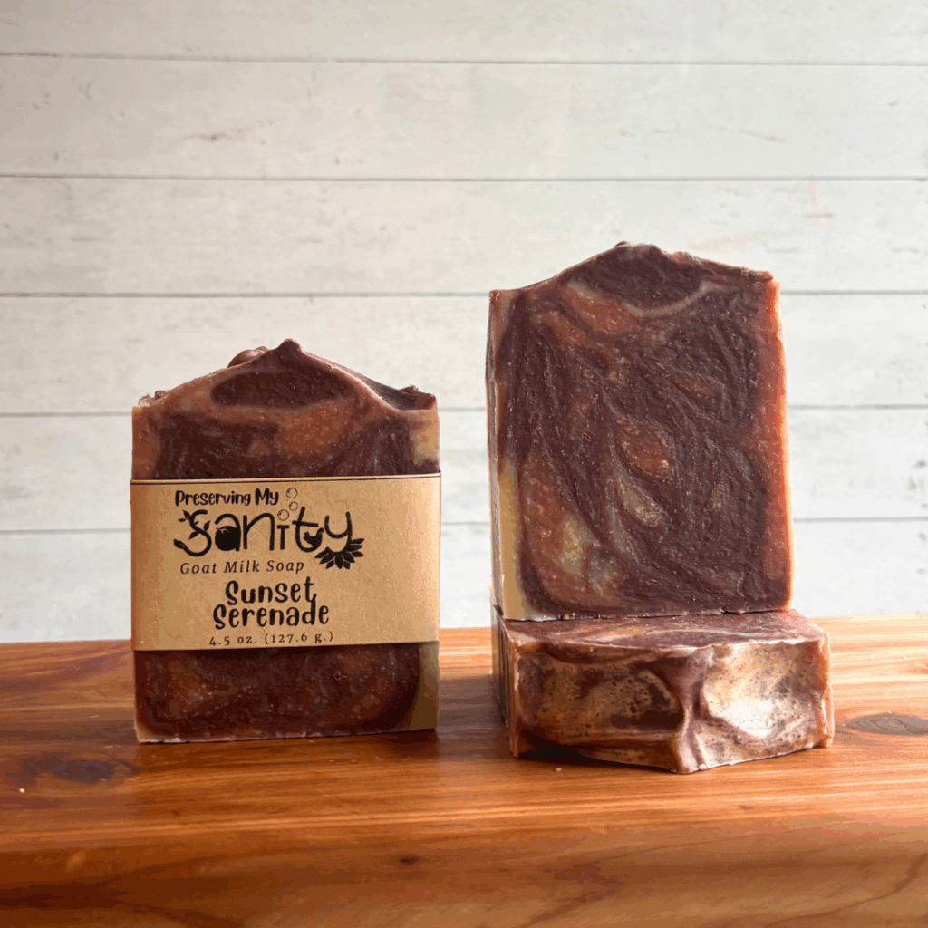Three bars of Sunset Serenade goat milk soap situated on a cedar bath board with a white paneled background. Soap is made with an essential oil blend, natural plant-based colorants, and locally sourced goat milk.