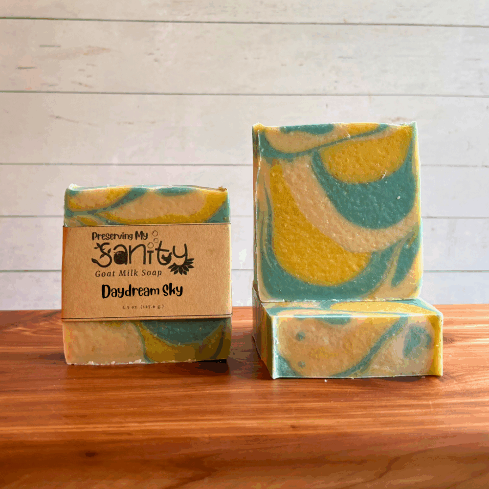 Three bars of Daydream Sky goat milk soap. The soap is a groovy swirl design of light blue, bright yellow, and white to mimic clouds and sky and sunshine. Two bars are displayed to show the design and top of the bars, while the third shows the label.