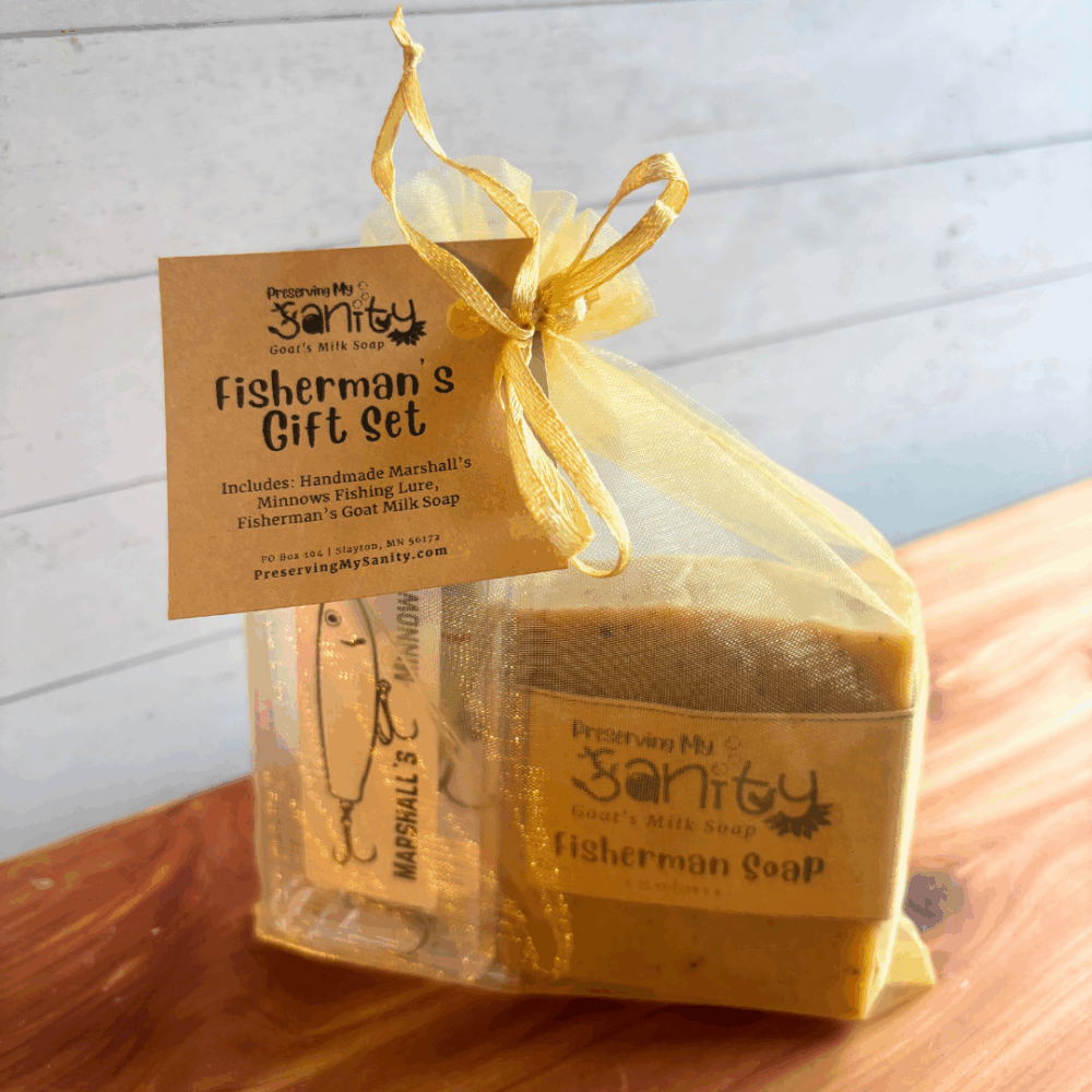 Fisherman’s Gift Set by Preserving My Sanity wrapped in a gold organza gift bag on a natural cedar wood surface with a white paneled background. This handmade gift set includes a 4.25-ounce bar of Fisherman’s goat milk soap scented with anise and lemon, along with a handmade fishing lure by Marshall’s Minnows packaged in a clear plastic lure case.