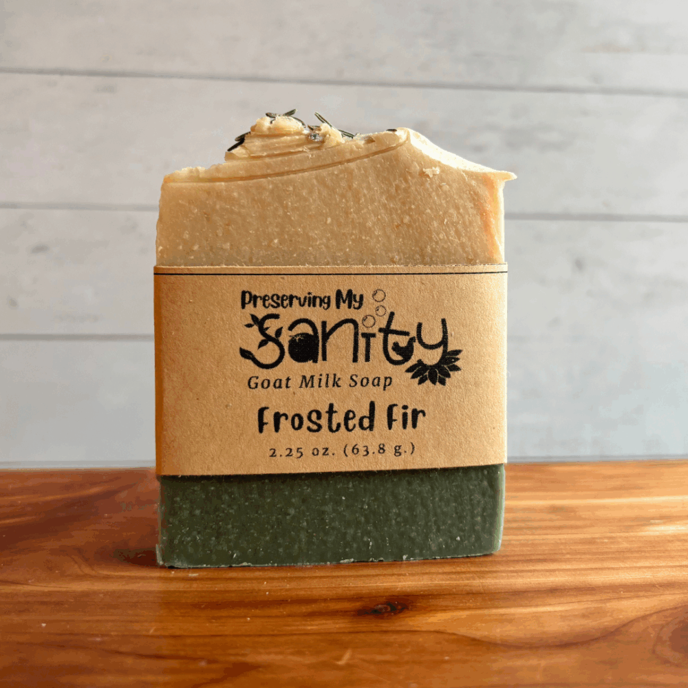 Single 2.25 ounce half-size bar of Frosted Fir goat milk soap by Preserving My Sanity displayed upright on a cedar bath board with a light paneled background. The handmade soap features creamy white and deep evergreen layers inspired by a festive holiday fragrance blend of wild berry, pineapple, pacific breeze, silver fir, blue spruce, plumeria, and musk.