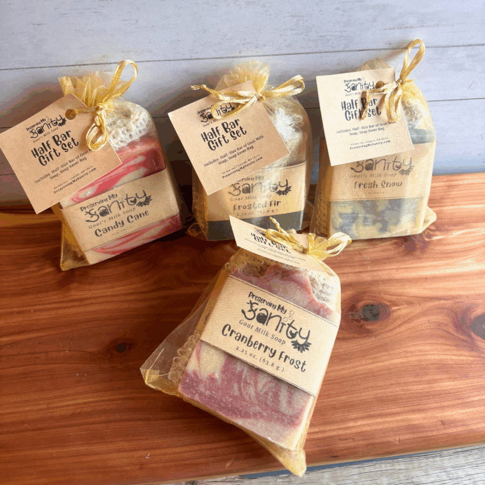 Collection of four holiday Half Bar Gift Sets by Preserving My Sanity displayed on a cedar bath board with a light paneled background. Each gift set includes a half-size goat milk soap bar and a natural sisal soap saver bag packaged in a sheer gold organza bag. Holiday scents featured include Candy Cane, Frosted Fir, Fresh Snow, and Cranberry Frost.