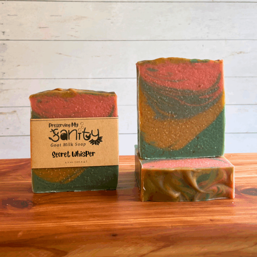 Three bars of Secret Whisper goat milk soap displayed in a row on a cedar bath board with a white paneled background. Two bars are displayed to show the swirly layers of forest green, gold, sea blue, and coral found in this unique paranormal themed goat milk soap. The third bar is laid flat on the board to display the textured and swirled soap top.