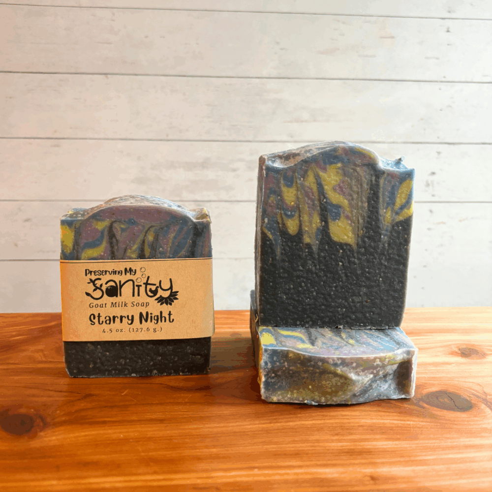 Three handcrafted bars of Starry Night goat milk soap displayed on a warm wood surface against a light paneled background. One labeled bar stands upright while two unlabeled bars showcase the deep charcoal base and swirling blue, gold, and purple details inspired by a secluded evening sky. The soap features a masculine blend of citrus, florals, woods, and musk, with notes of bergamot, grapefruit, lavender, sandalwood, vanilla, and amber.