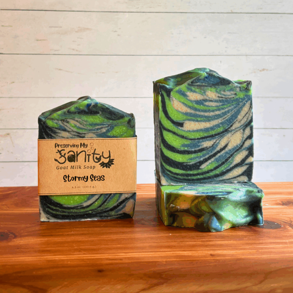 Three bars of Stormy Seas goat milk soap displayed on a cedar bath board with a white paneled background. Two bars are displayed to show the wispy swirls of black, blue, lime green, and white in this paranormal-themed soap. The third bar is laying flat on the board to display the textured soap top.