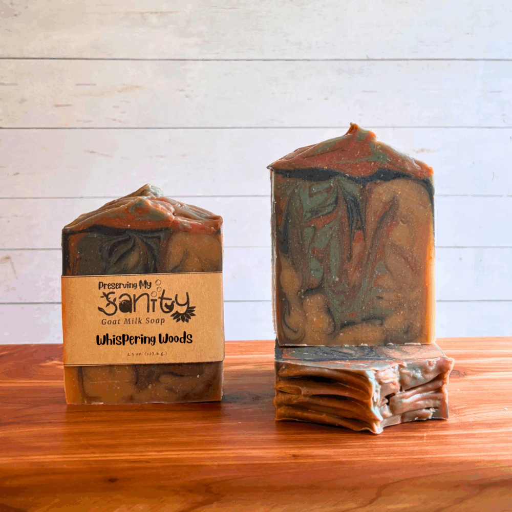 Three bars of Whispering Woods goat milk soap. This soap has an eerie blend of light brown, dark brown, green, and black swirls that are perfect for the spooky season. Two bars are displayed to show the design and top of the bars, while the third shows the label.