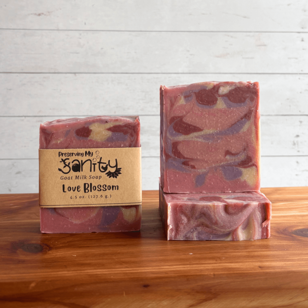 Three bars of Love Blossom goat milk soap displayed on a cedar board against a white shiplap background. The center bar stands upright with the label visible, while the other two bars showcase soft swirls of mauve, blush pink, cream, and lavender throughout the soap.