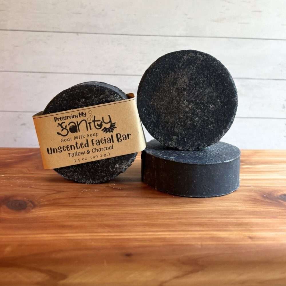 Three round Unscented Charcoal Facial Soap bars displayed on a cedar bath board against a white paneled background. The center bar features the kraft label, while the two unlabeled bars showcase the smooth, matte black finish created by activated charcoal. One bar stands upright to display its full round shape, and another is laid flat to highlight the bar's thickness, emphasizing the simple, handcrafted design of this fragrance-free facial soap.