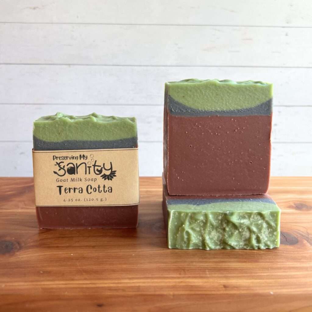 Three bars of Terra Cotta handmade goat milk soap displayed on a cedar bath board against a white paneled background. The soap features earthy layers of terracotta brown, charcoal gray, and soft sage green inspired by natural clay and calming spa tones. One labeled bar and one naked bar are displayed upright to showcase the layered design, while the third bar is laid flat on the board to highlight the textured top of the soap.