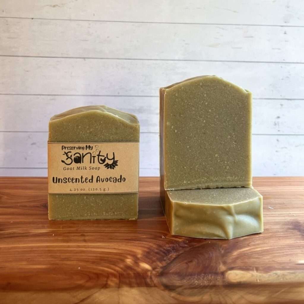 Two upright bars and one flat bar of Preserving My Sanity Unscented Avocado Soap are displayed on a warm cedar bath board against a white paneled background. The bar on the left is wrapped with a kraft paper label reading "Unscented Avocado," while the unwrapped bar on the right highlights the soap's soft green color and smooth, minimalist finish. A third bar lies flat beneath it, showcasing the top edge and handcrafted shape of this fragrance-free soap made with real avocado and avocado oil.