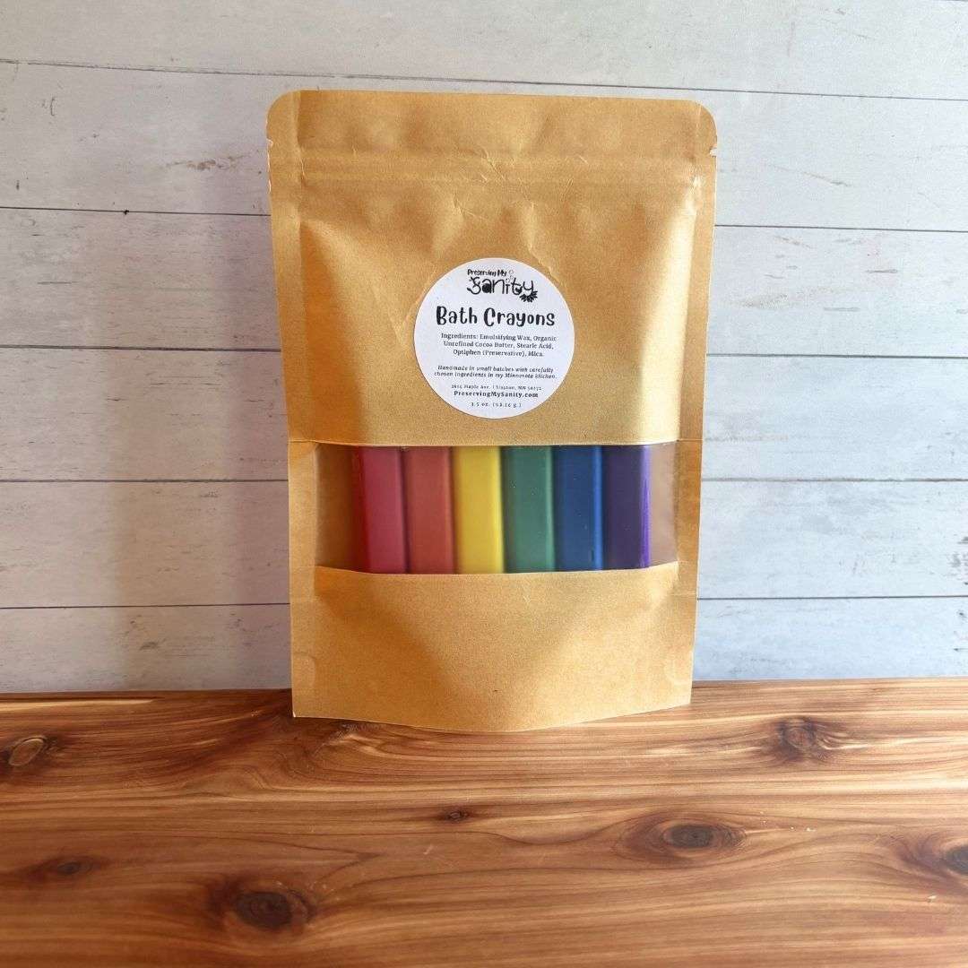 Bath Crayons packaged in a kraft stand-up pouch with a clear window, displayed on a cedar bath board against a white paneled background. The window reveals six colorful bath crayons in red, orange, yellow, green, blue, and purple, while a round label on the front identifies the product.