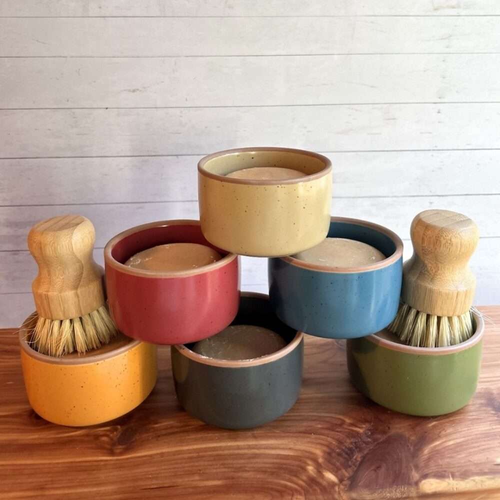 Seven handmade goat milk dish soap sets are stacked in a pyramid display on a cedar bath board against a white paneled background. Each set features a round bar of dish soap in a colorful ceramic ramekin from Mora Ceramics, with ramekins shown in cream, coral, blue, gray, olive green, and mustard yellow. Two sets include bamboo-handled dish brushes with natural sisal bristles resting inside the ramekins, highlighting the reusable kitchen cleaning set.