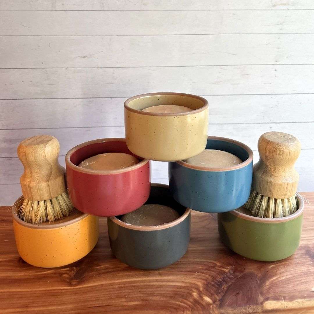 Seven handmade goat milk dish soap sets are stacked in a pyramid display on a cedar bath board against a white paneled background. Each set features a round bar of dish soap in a colorful ceramic ramekin from Mora Ceramics, with ramekins shown in cream, coral, blue, gray, olive green, and mustard yellow. Two sets include bamboo-handled dish brushes with natural sisal bristles resting inside the ramekins, highlighting the reusable kitchen cleaning set.