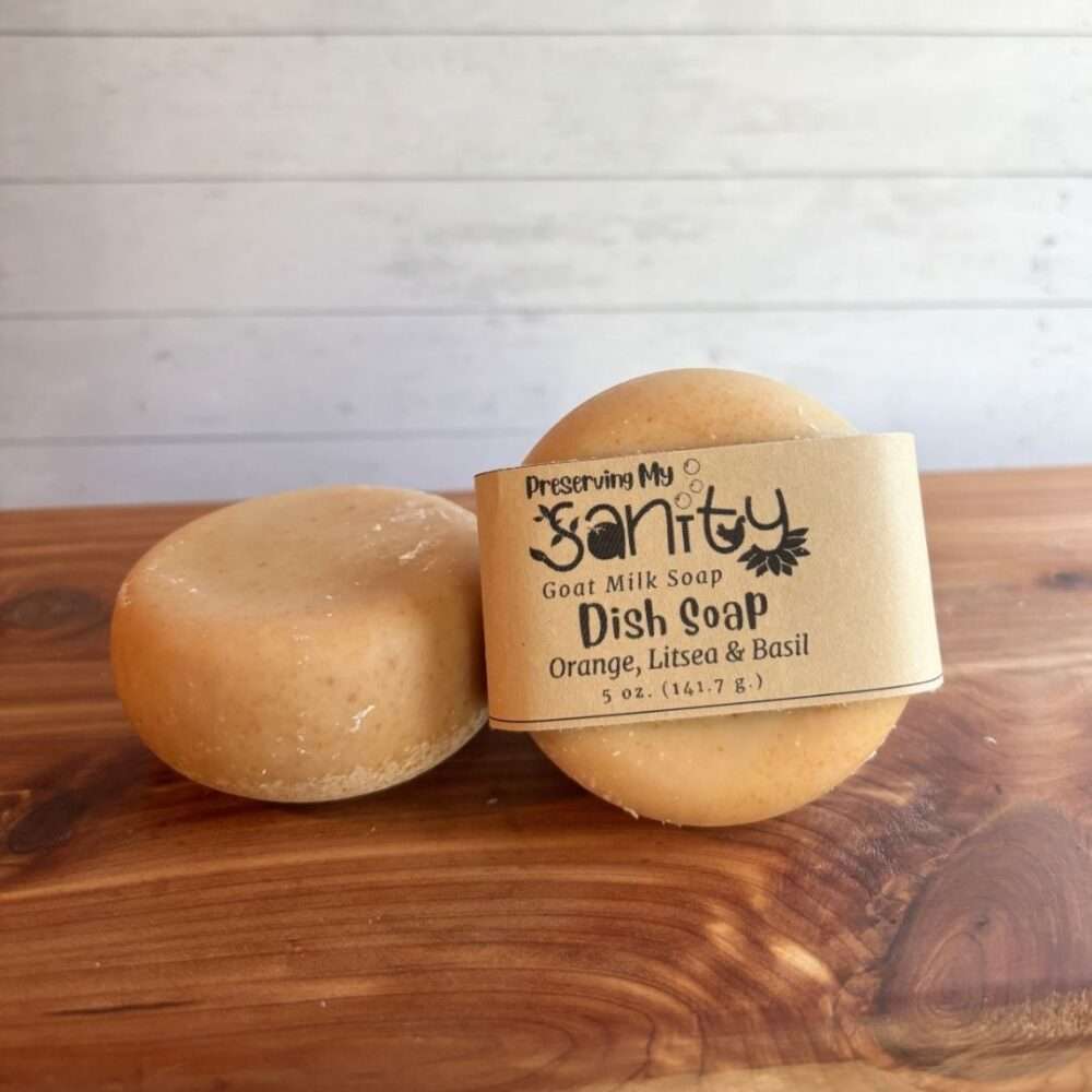 Two round bars of handmade goat milk dish soap are displayed on a cedar bath board against a white paneled background. One bar is wrapped in a kraft paper label reading "Dish Soap, Orange, Litsea & Basil," while the second unwrapped bar rests beside it, showing the smooth, rounded shape and natural golden color of the soap.