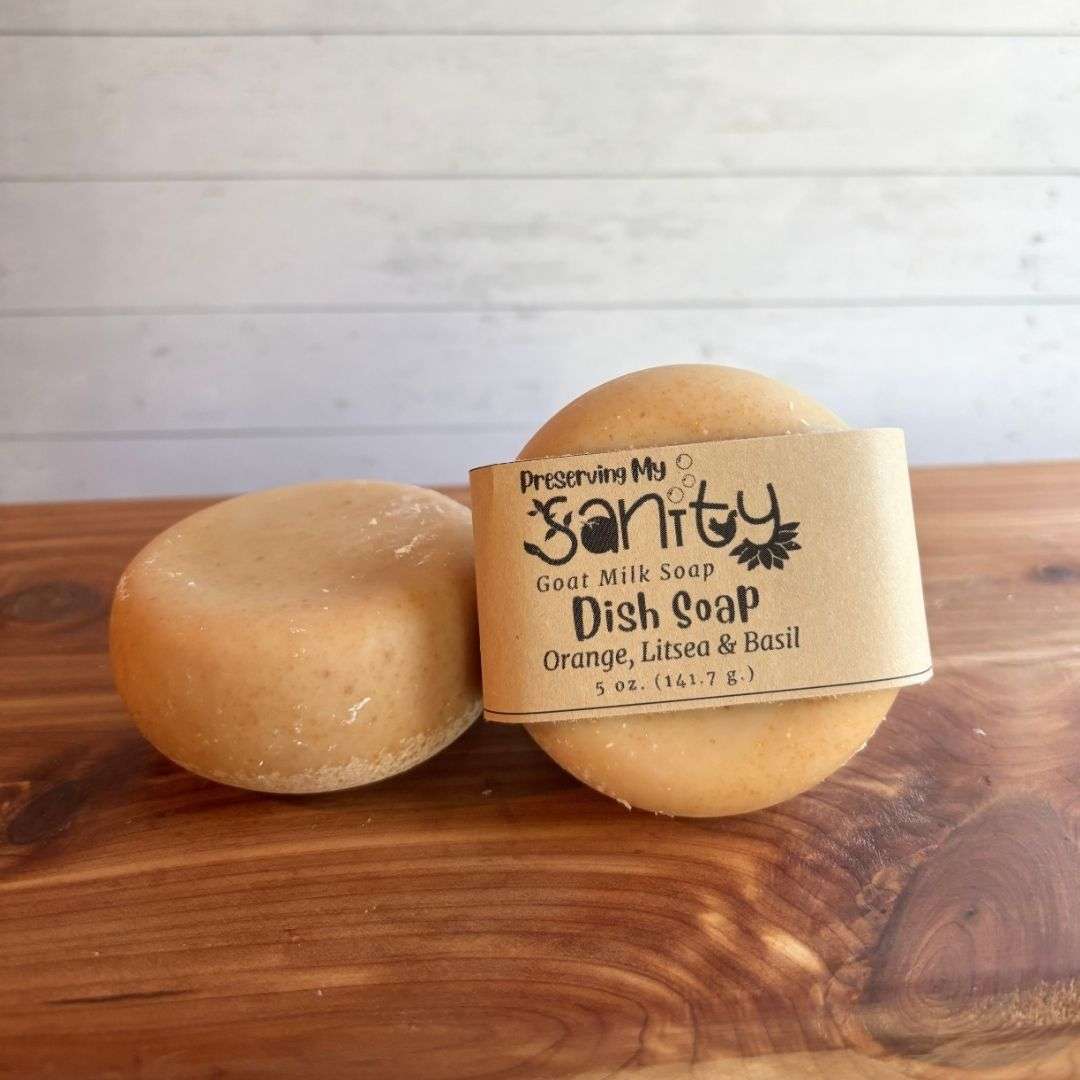 Two round bars of handmade goat milk dish soap are displayed on a cedar bath board against a white paneled background. One bar is wrapped in a kraft paper label reading "Dish Soap, Orange, Litsea & Basil," while the second unwrapped bar rests beside it, showing the smooth, rounded shape and natural golden color of the soap.