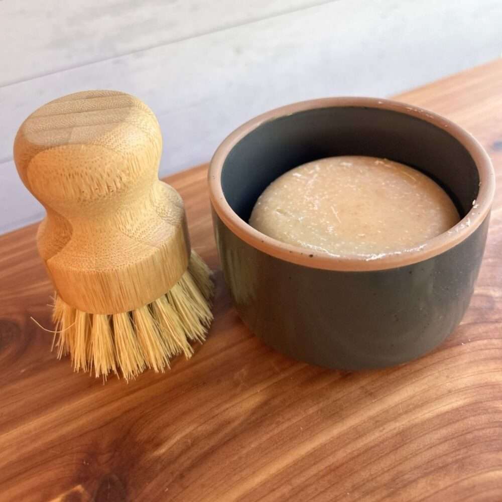 A handmade goat milk dish soap set is displayed on a cedar bath board against a white paneled background. The set includes a round bar of dish soap nestled inside a charcoal gray ceramic ramekin from Mora Ceramics, paired with a bamboo-handled dish brush featuring natural sisal bristles.