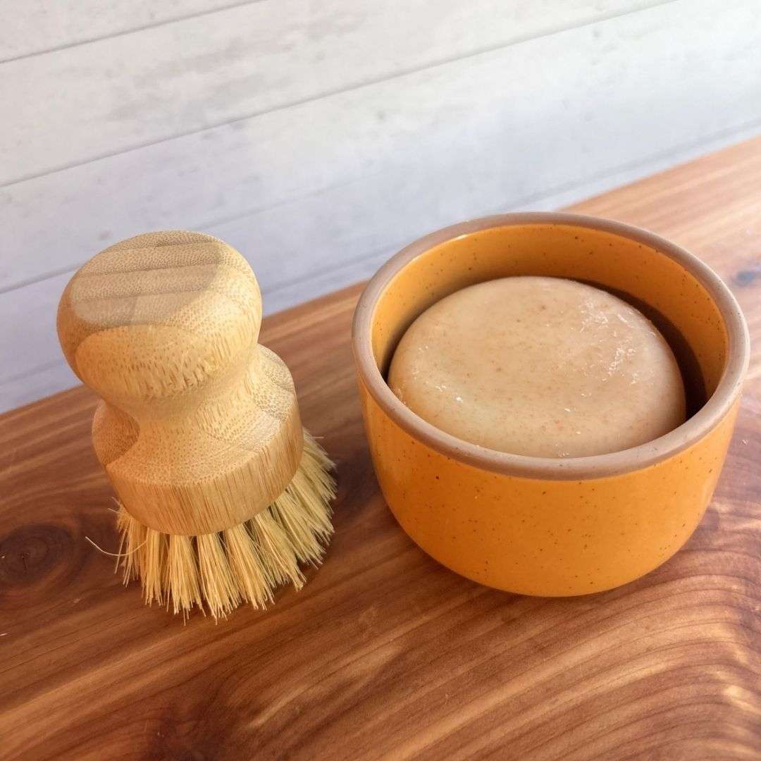 A handmade goat milk dish soap set is displayed on a cedar bath board against a white paneled background. The set includes a round bar of dish soap nestled inside a yellow ceramic ramekin from Mora Ceramics, paired with a bamboo-handled dish brush featuring natural sisal bristles.