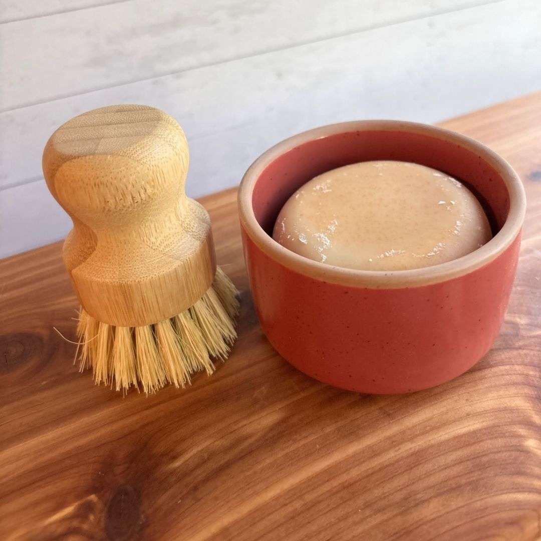 A handmade goat milk dish soap set is displayed on a cedar bath board against a white paneled background. The set includes a round bar of dish soap nestled inside a red ceramic ramekin from Mora Ceramics, paired with a bamboo-handled dish brush featuring natural sisal bristles.
