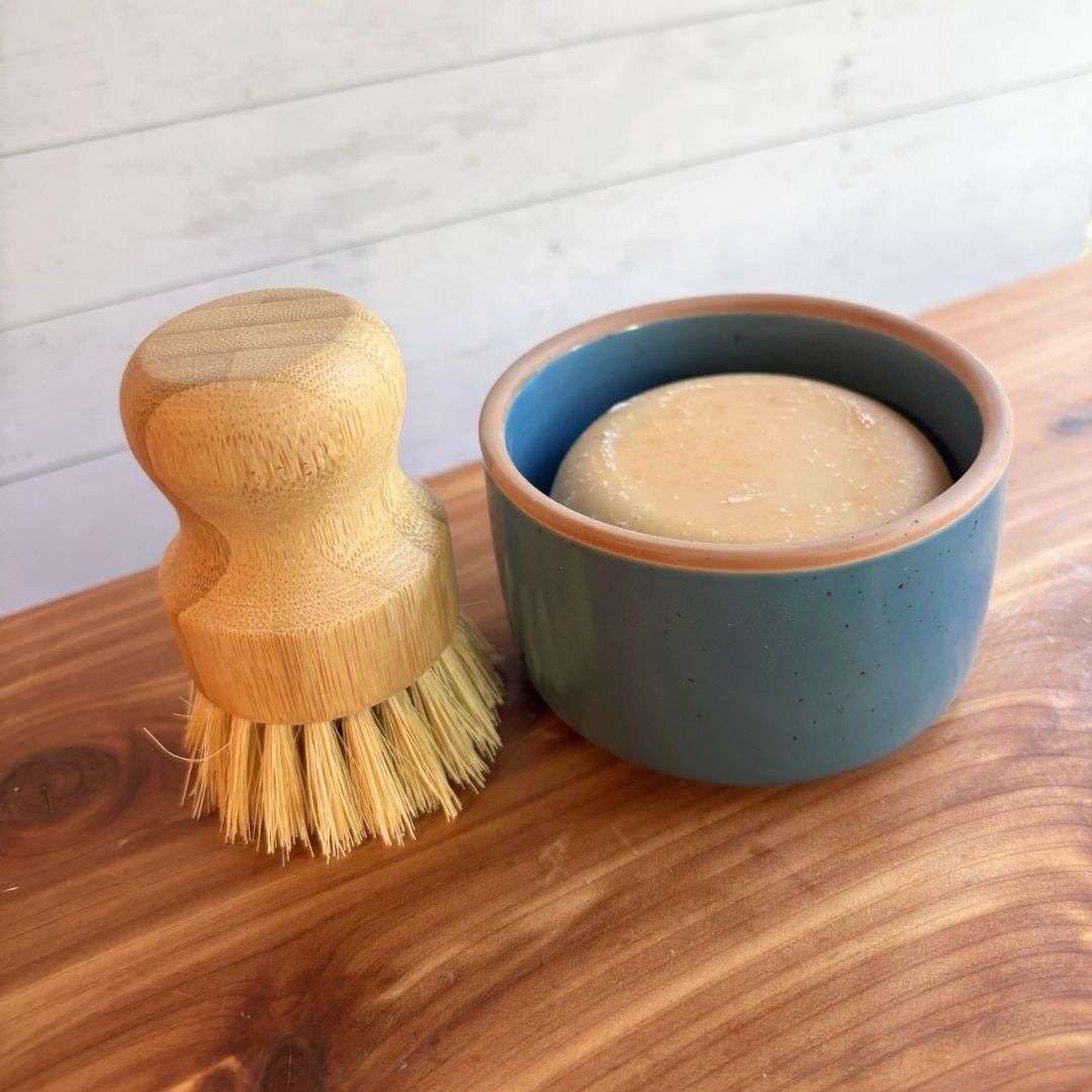 A handmade goat milk dish soap set is displayed on a cedar bath board against a white paneled background. The set includes a round bar of dish soap nestled inside a blue ceramic ramekin from Mora Ceramics, paired with a bamboo-handled dish brush featuring natural sisal bristles.