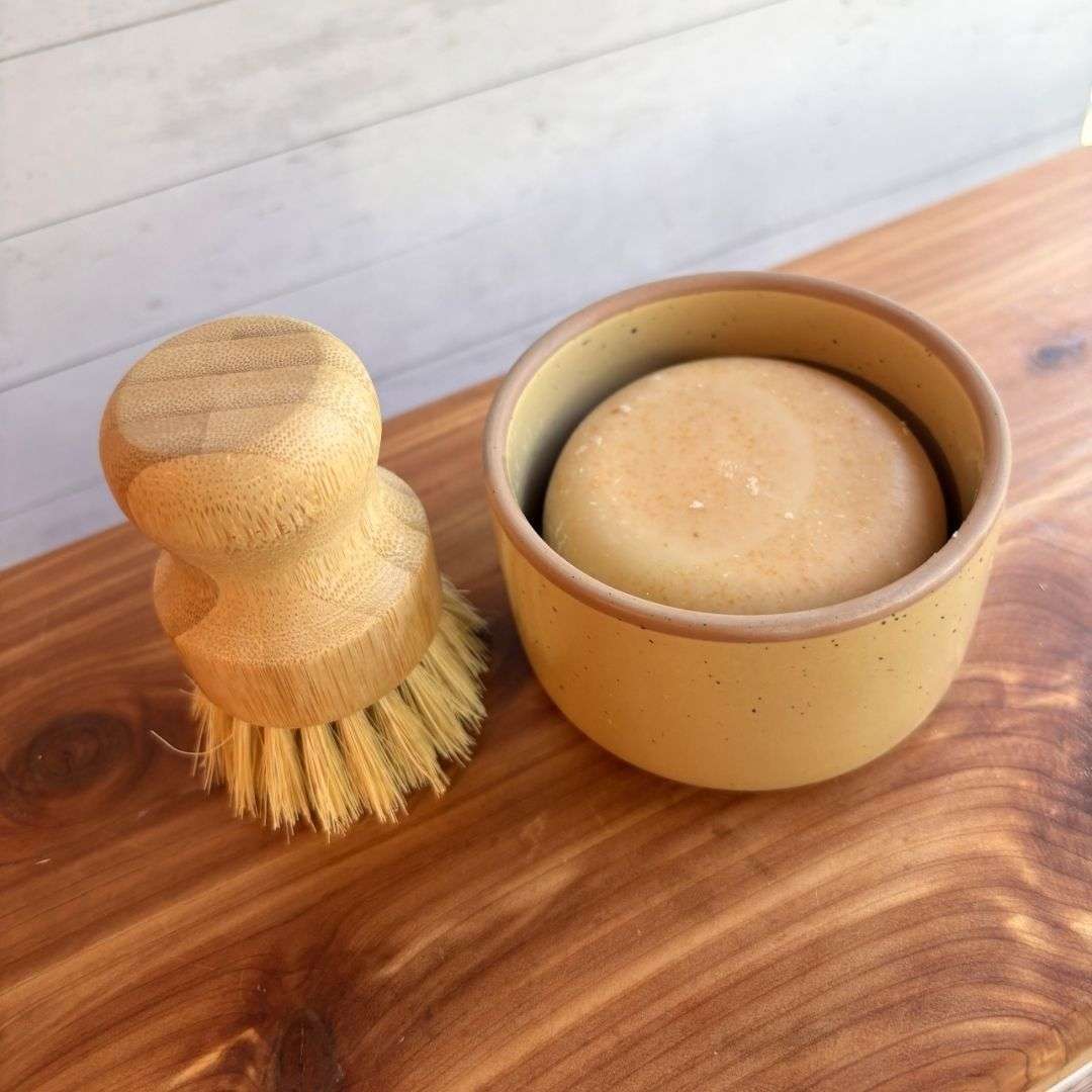 A handmade goat milk dish soap set is displayed on a cedar bath board against a white paneled background. The set includes a round bar of dish soap nestled inside a biscuit ceramic ramekin from Mora Ceramics, paired with a bamboo-handled dish brush featuring natural sisal bristles.