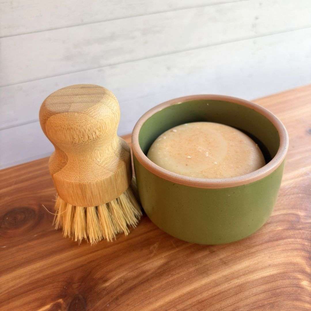 A handmade goat milk dish soap set is displayed on a cedar bath board against a white paneled background. The set includes a round bar of dish soap nestled inside a green ceramic ramekin from Mora Ceramics, paired with a bamboo-handled dish brush featuring natural sisal bristles.