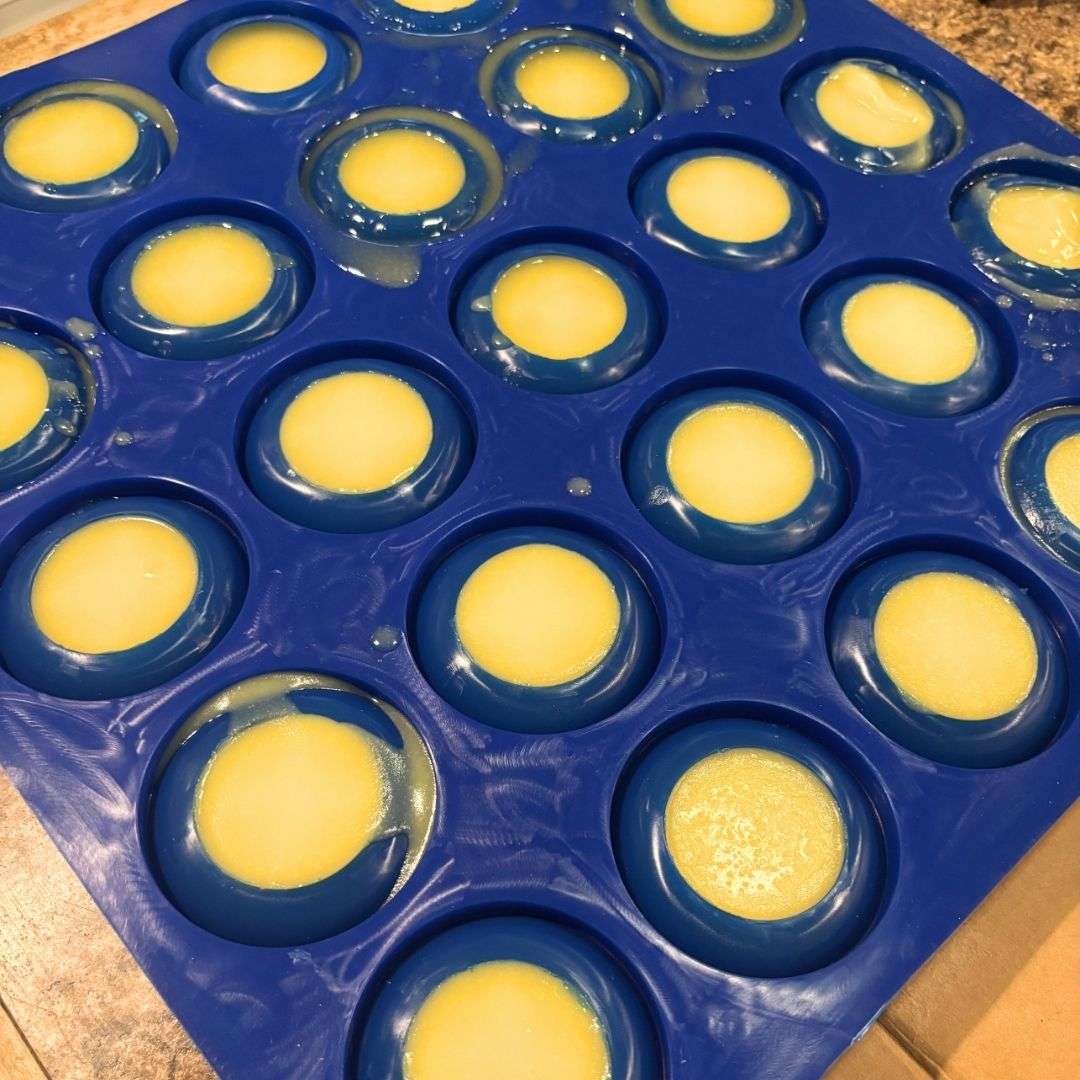 Process photo of handmade goat milk dish soap in a blue silicone mold with multiple round cavities. Each cavity is filled with creamy golden dish soap that has been poured and is beginning to set, showing the soap before it is cured.