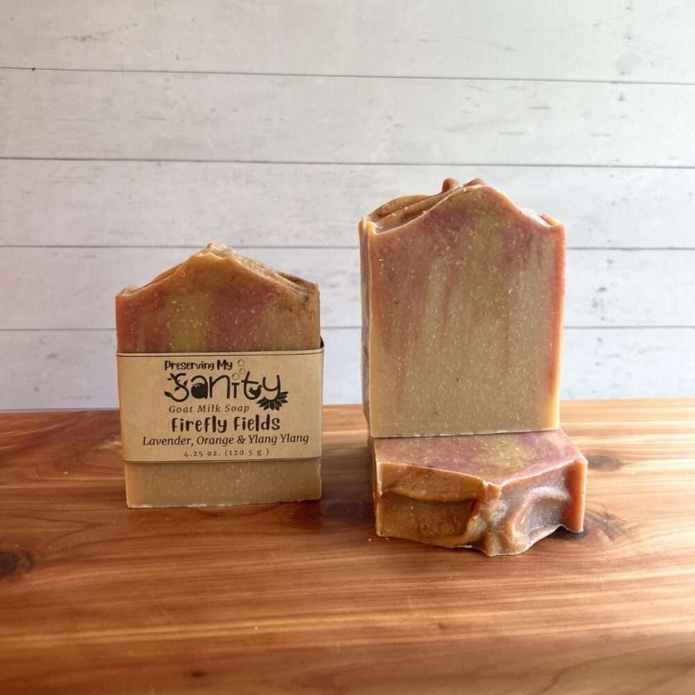 Three bars of Firefly Fields goat milk soap displayed on a cedar bath board against a white paneled background. One bar stands upright with the signature kraft label, one stands upright without a label to showcase the warm golden and soft mauve swirls, and one bar lies flat in front to highlight the textured top. Handmade with lavender, orange, and ylang ylang essential oils, the soap features a soft, earthy color palette inspired by glowing summer evenings.