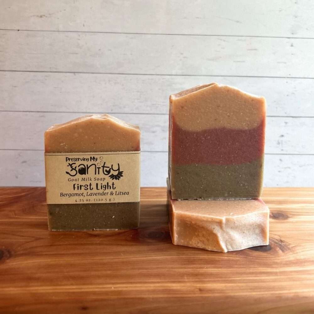 Three bars of First Light goat milk soap displayed on a cedar bath board against a white shiplap background. One bar stands upright with the signature kraft label, one stands upright without a label to showcase the clean horizontal layers of soft peach, muted rose, and earthy olive green, and one bar lies flat in front to highlight the smooth top. Scented with bergamot, lavender, and litsea, this handcrafted soap is inspired by the warm colors of the first light of day.