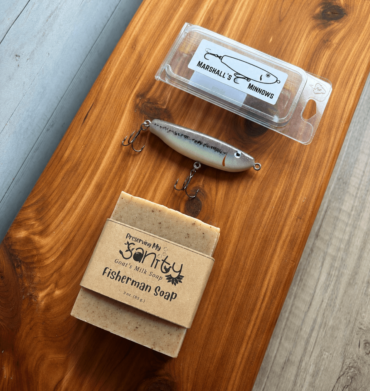 Fisherman's Gift Set displayed on a cedar bath board. The set includes a 3-ounce bar of Fisherman's Goat Milk Soap wrapped in the signature kraft label, a handmade Marshall's Minnows fishing lure with a black-and-white finish, and the lure's clear protective storage case.