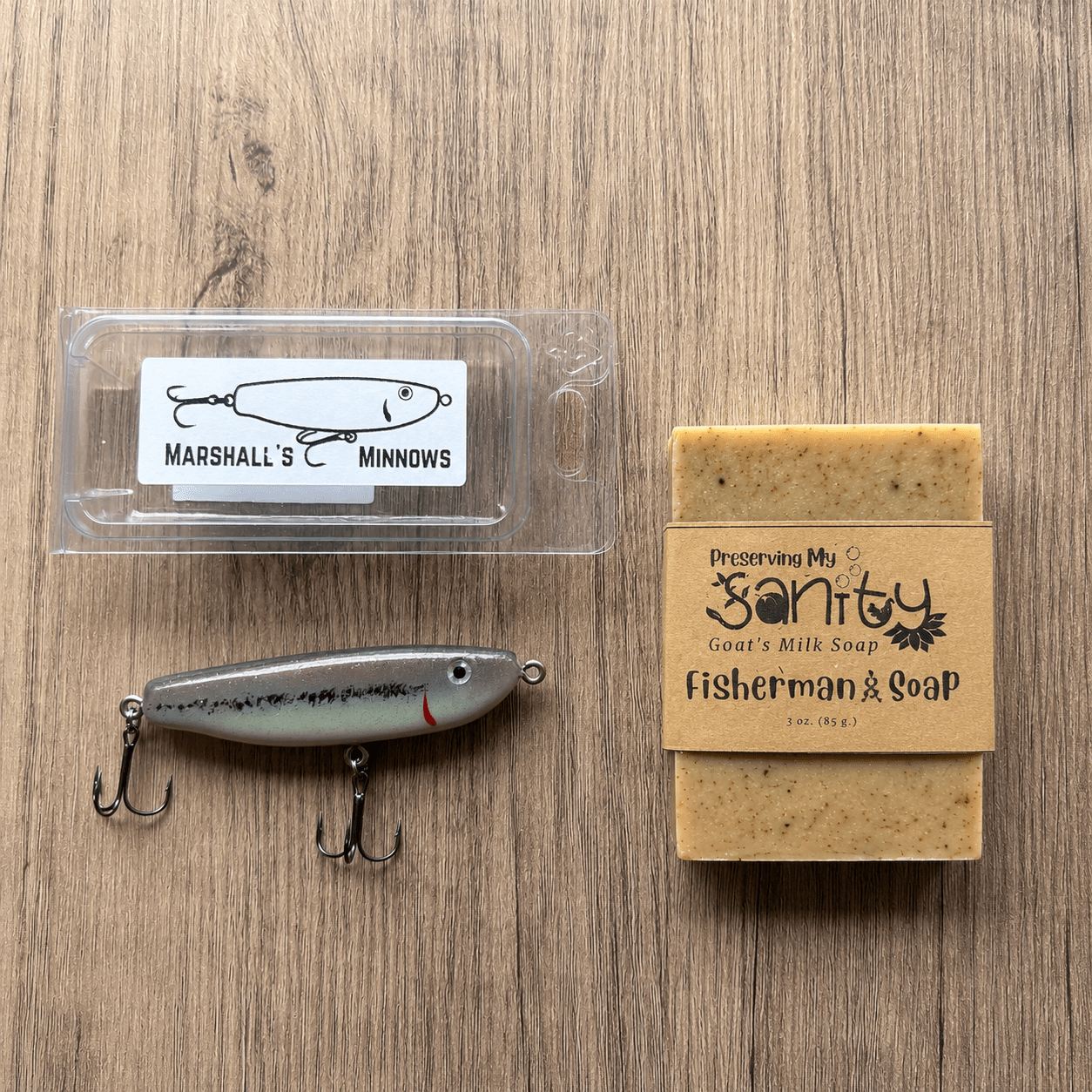 Fisherman's Gift Set components displayed on a light wood surface. The arrangement includes a 3-ounce bar of Fisherman's Goat Milk Soap wrapped in the signature kraft label, a handmade Marshall's Minnows fishing lure with a black-and-white finish, and the lure's clear protective storage case.