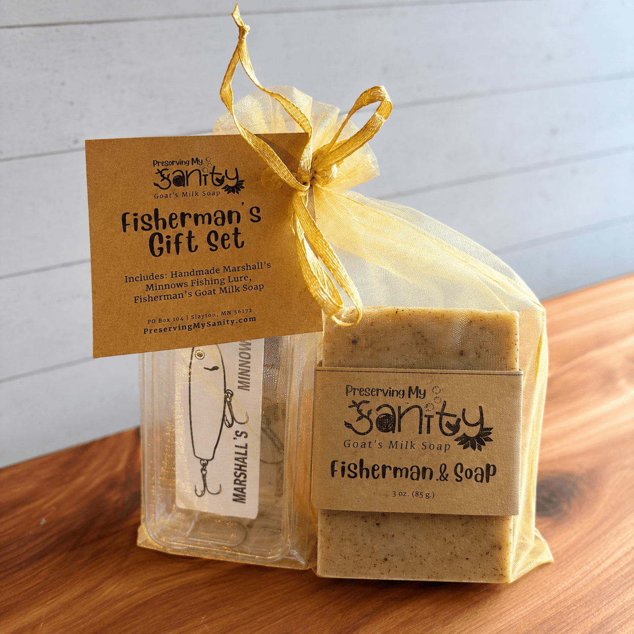Fisherman's Gift Set displayed on a cedar bath board against a white paneled background. The gift set is packaged in a sheer gold organza bag tied with a gold ribbon and includes a 3-ounce bar of Fisherman's Goat Milk Soap wrapped in the signature kraft label alongside a handmade fishing lure in its clear protective case.