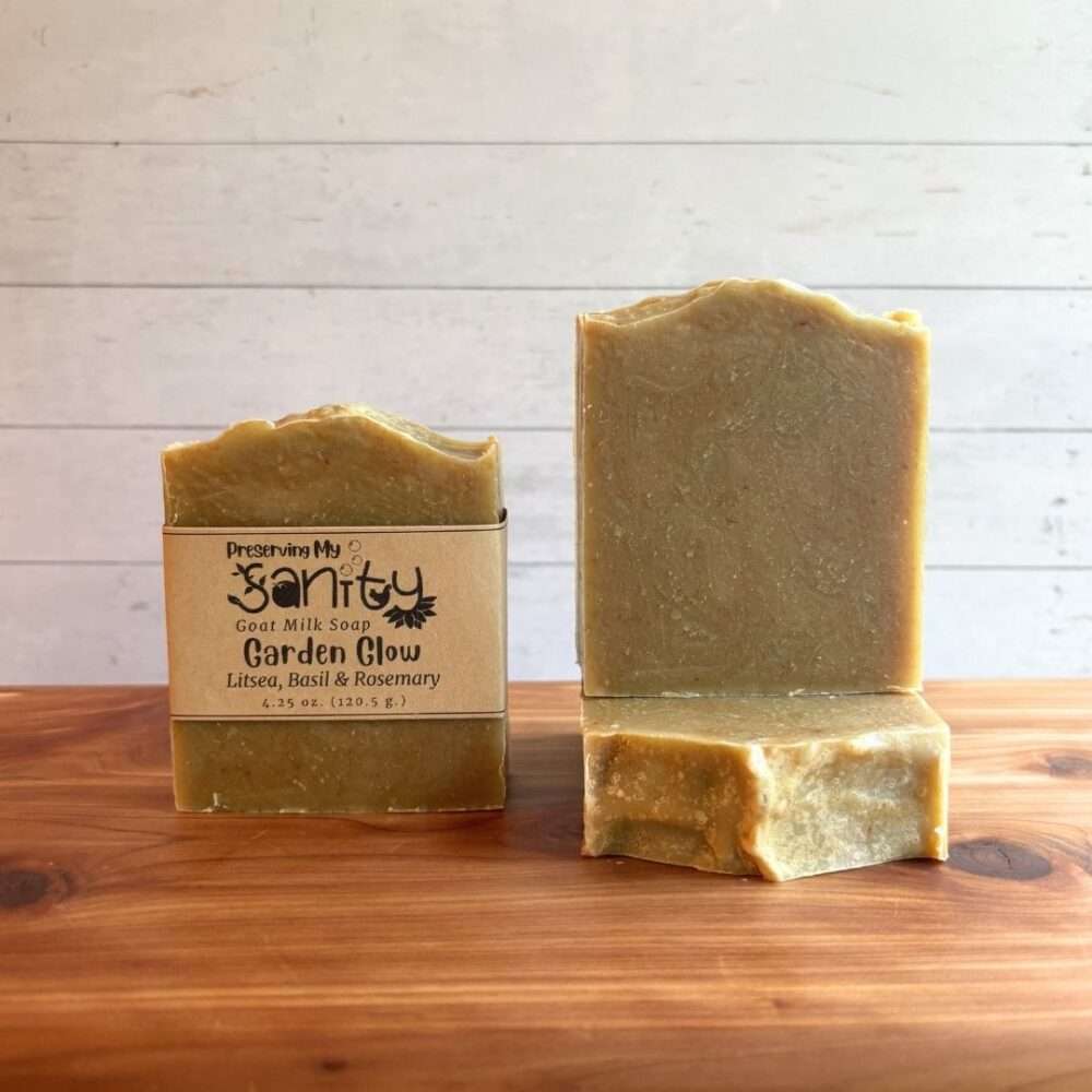 Three bars of Garden Glow goat milk soap displayed on a cedar bath board against a white shiplap background. One bar stands upright with the signature kraft label, one stands upright without a label to showcase its soft olive-green color, and one bar lies flat in front to reveal the smooth top. Scented with litsea, basil, and rosemary essential oils, this handcrafted soap has a simple, natural appearance that reflects its fresh herbal fragrance.