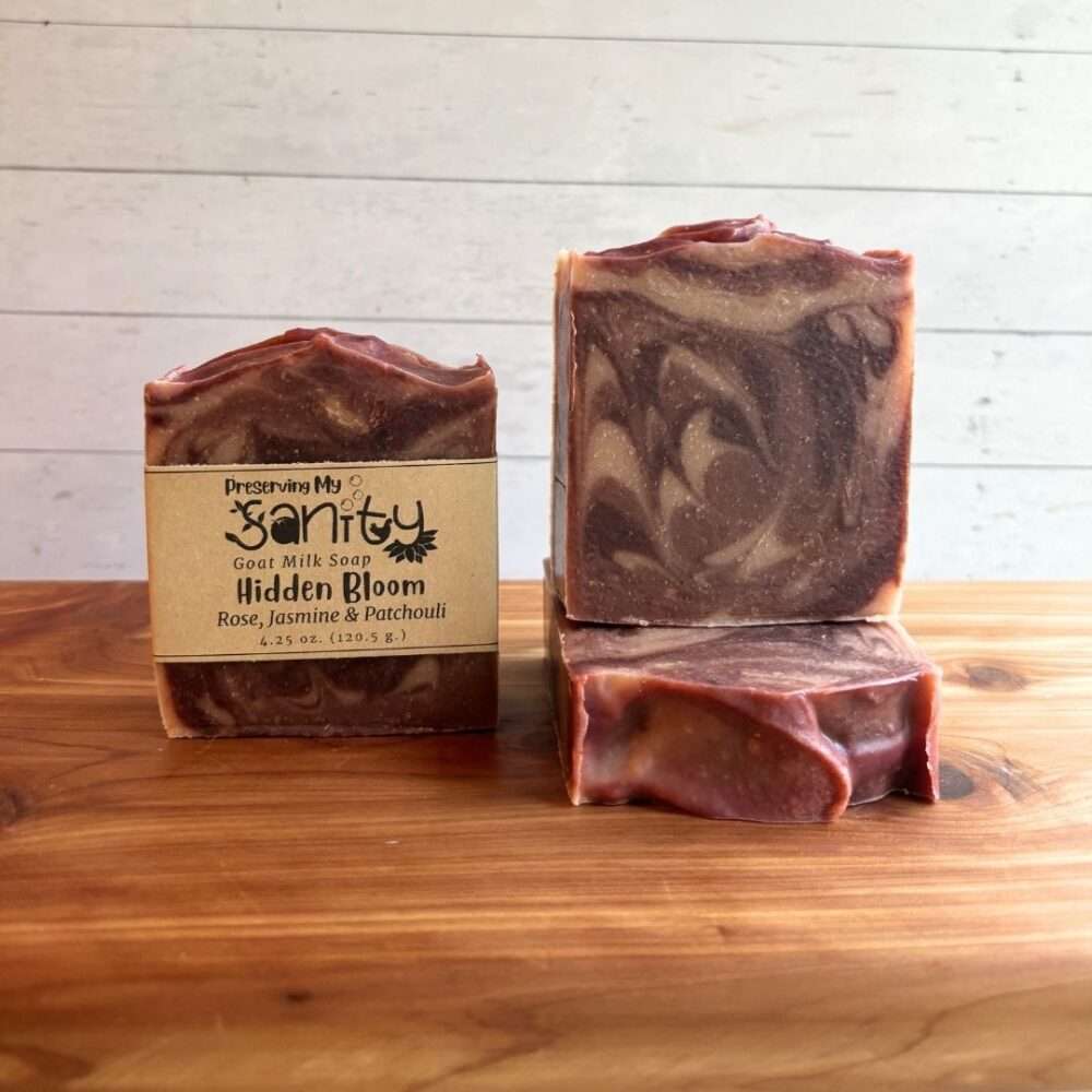 Three bars of Hidden Bloom goat milk soap displayed on a cedar bath board against a white shiplap background. One bar stands upright with the signature kraft label, one stands upright without a label to showcase the rich marbled swirls of plum, mauve, and creamy beige, and one bar lies flat in front to reveal the textured top. Scented with rose, jasmine, and patchouli, this handcrafted soap features an elegant floral-inspired design with warm, earthy tones.