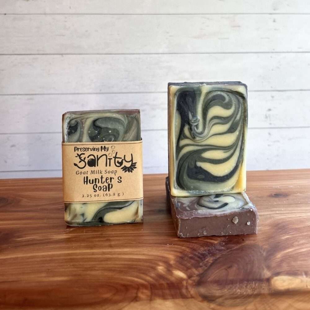 Three bars of Hunter's Soap are displayed on a warm wooden surface against a white shiplap background. On the left, a labeled 2.25 oz bar showcases striking swirls of charcoal black, sage green, cream, and earthy brown. On the right, an unlabeled upright bar highlights the bold marbled swirl pattern, while a third bar lies flat beneath it, revealing the soap's top.