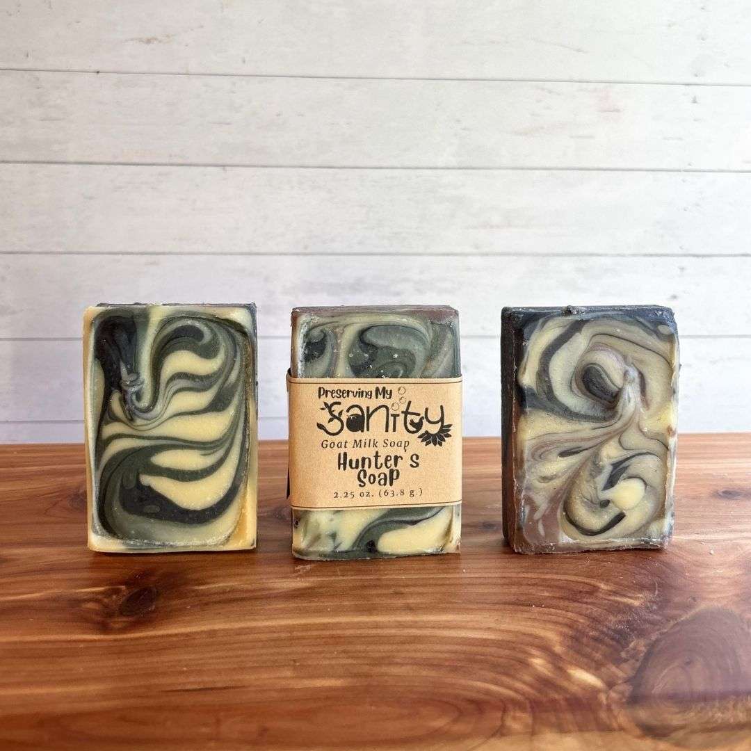 Three bars of Hunter's Soap arranged side by side on a cedar bath board against a white shiplap background. The center bar is wrapped with the signature Preserving My Sanity kraft label, while the bars on the left and right are unwrapped to showcase the striking marbled swirls of charcoal black, sage green, cream, and earthy brown.