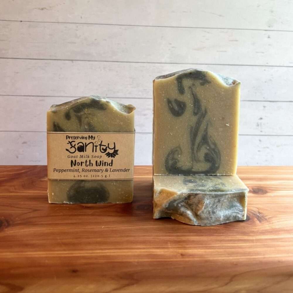 Three bars of North Wind goat milk soap are displayed on a cedar bath board against a white shiplap background. One bar stands upright with the signature kraft label, one stands upright without a label to showcase its soft sage-green base with wispy charcoal swirls, and one bar lies flat in front to reveal the textured top. Scented with peppermint, rosemary, and lavender essential oils, this handcrafted soap has a clean, nature-inspired design that reflects its fresh herbal fragrance.