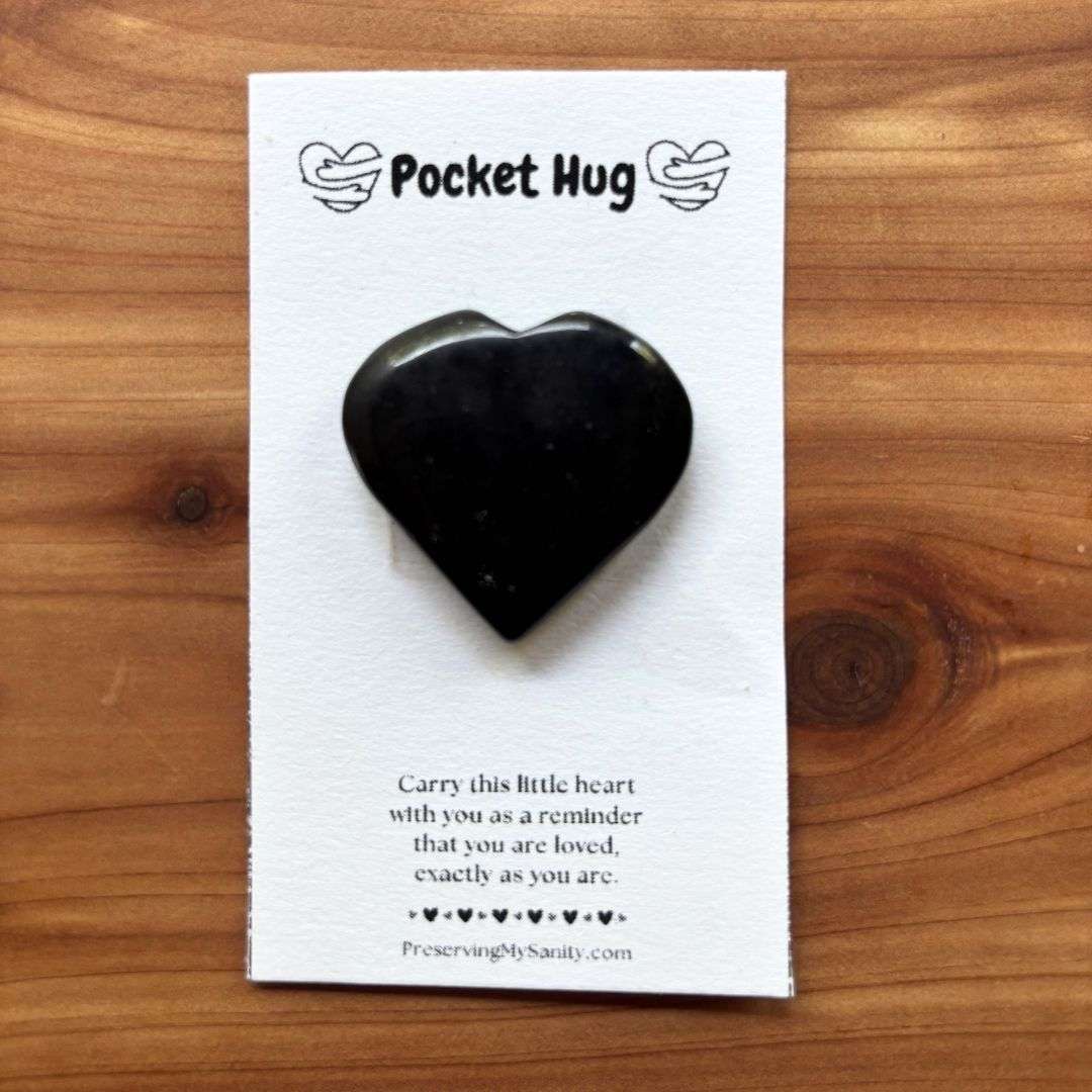 A polished heart-shaped black tourmaline is mounted on a white "Pocket Hug" card and displayed on a warm cedar wood background. The card features the words "Pocket Hug" at the top with heart illustrations and a heartfelt message below: "Carry this little heart with you as a reminder that you are loved, exactly as you are." Small black hearts decorate the bottom above the Preserving My Sanity website, making this a thoughtful keepsake for encouragement, comfort, or gifting.