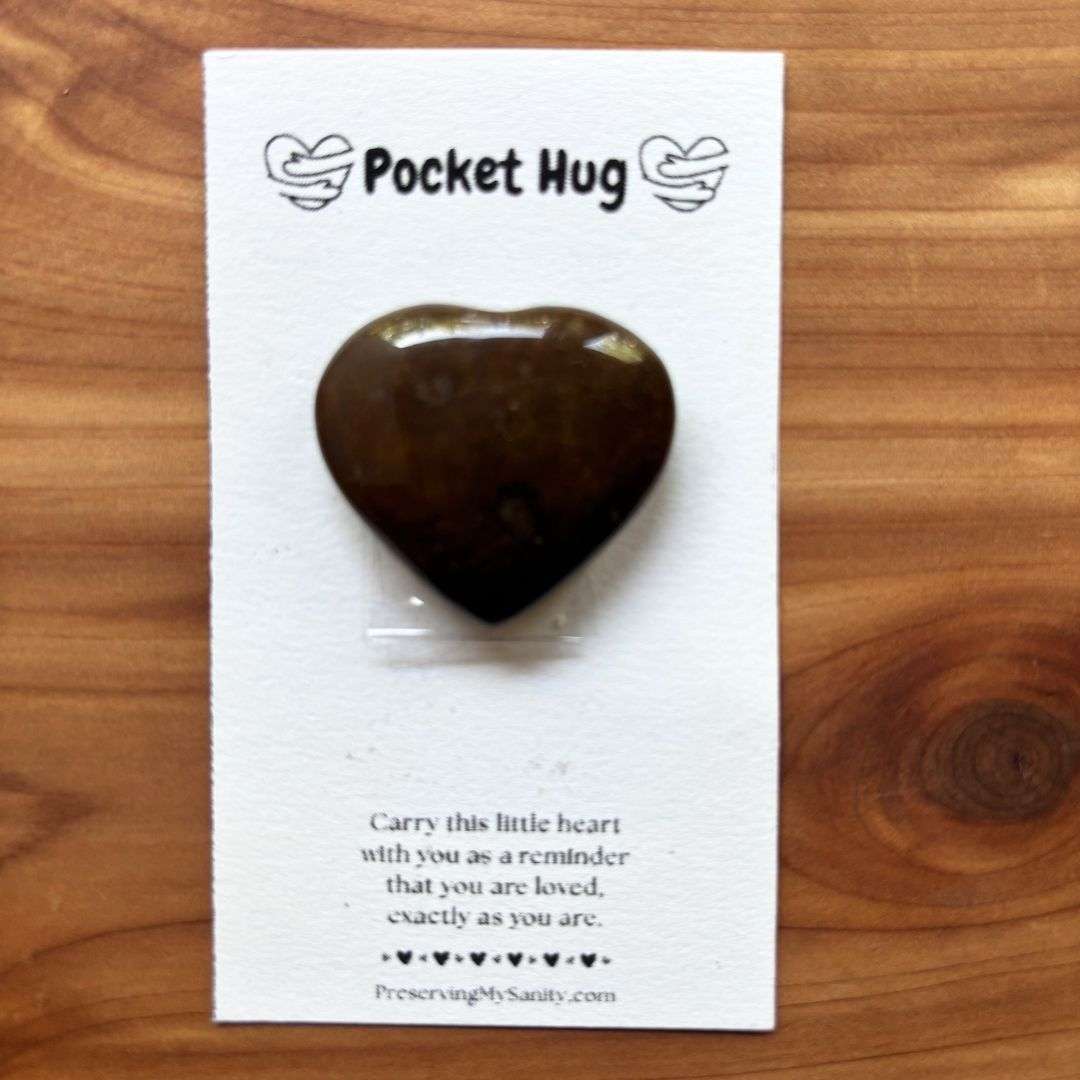 A polished heart-shaped fancy jasper is mounted on a white "Pocket Hug" card and displayed on a warm cedar wood background. The card features the words "Pocket Hug" at the top with heart illustrations and a heartfelt message below: "Carry this little heart with you as a reminder that you are loved, exactly as you are." Small black hearts decorate the bottom above the Preserving My Sanity website, making this a thoughtful keepsake for encouragement, comfort, or gifting.