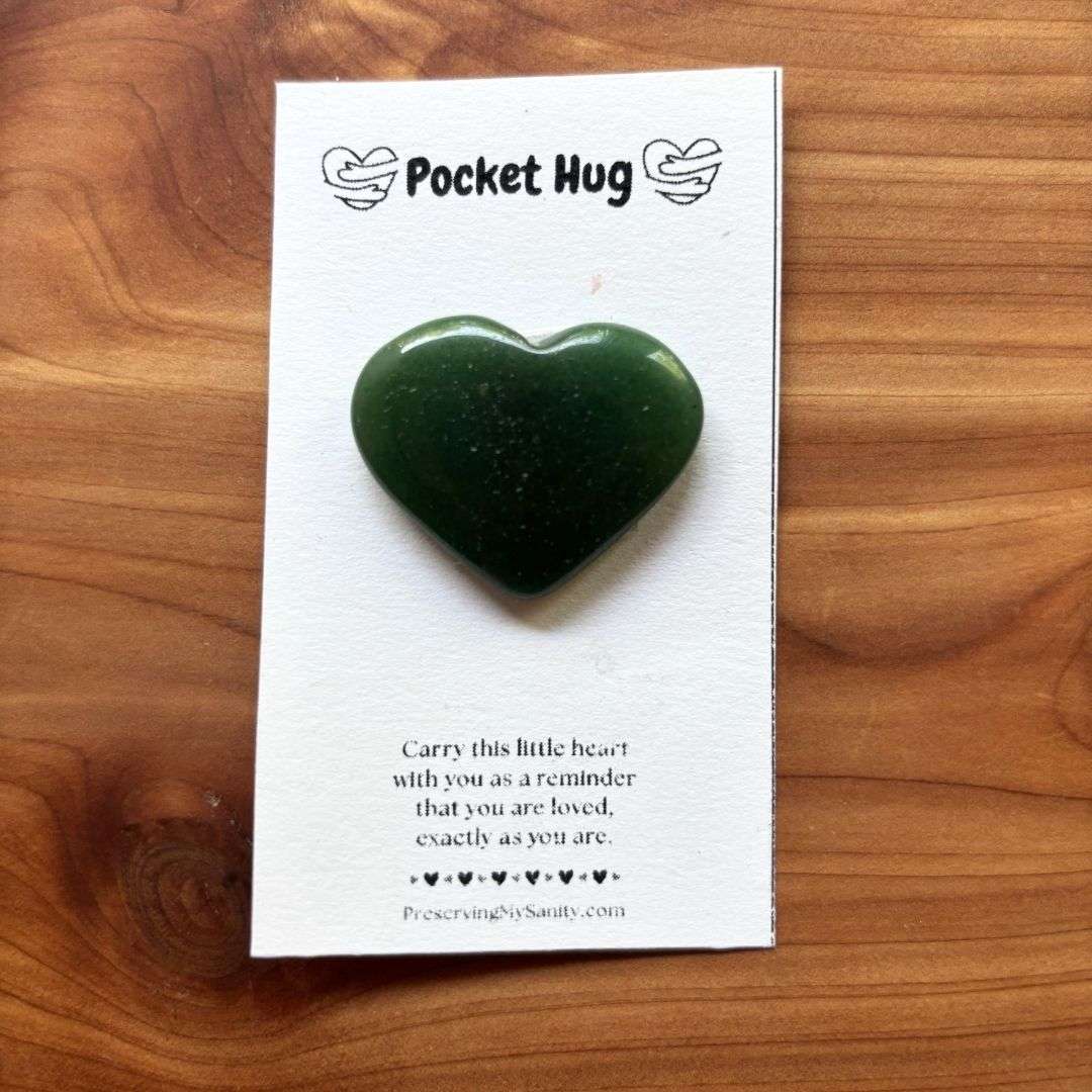 A polished heart-shaped green dark aventurine is mounted on a white "Pocket Hug" card and displayed on a warm cedar wood background. The card features the words "Pocket Hug" at the top with heart illustrations and a heartfelt message below: "Carry this little heart with you as a reminder that you are loved, exactly as you are." Small black hearts decorate the bottom above the Preserving My Sanity website, making this a thoughtful keepsake for encouragement, comfort, or gifting.