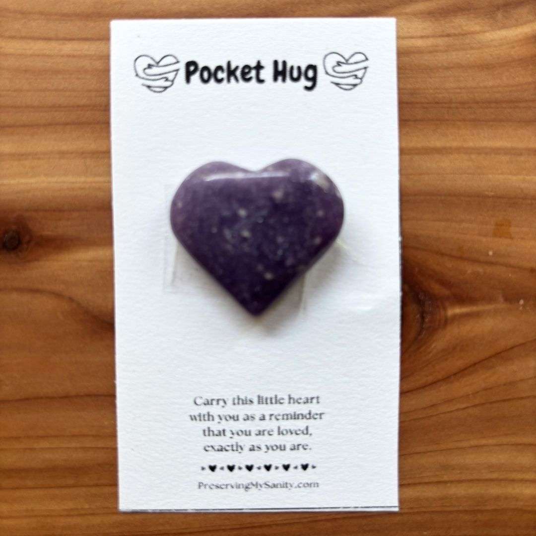 A polished heart-shaped lepidolite is mounted on a white "Pocket Hug" card and displayed on a warm cedar wood background. The card features the words "Pocket Hug" at the top with heart illustrations and a heartfelt message below: "Carry this little heart with you as a reminder that you are loved, exactly as you are." Small black hearts decorate the bottom above the Preserving My Sanity website, making this a thoughtful keepsake for encouragement, comfort, or gifting.