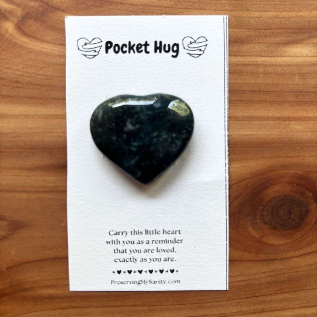 A polished heart-shaped moss agate is mounted on a white "Pocket Hug" card and displayed on a warm cedar wood background. The card features the words "Pocket Hug" at the top with heart illustrations and a heartfelt message below: "Carry this little heart with you as a reminder that you are loved, exactly as you are." Small black hearts decorate the bottom above the Preserving My Sanity website, making this a thoughtful keepsake for encouragement, comfort, or gifting.
